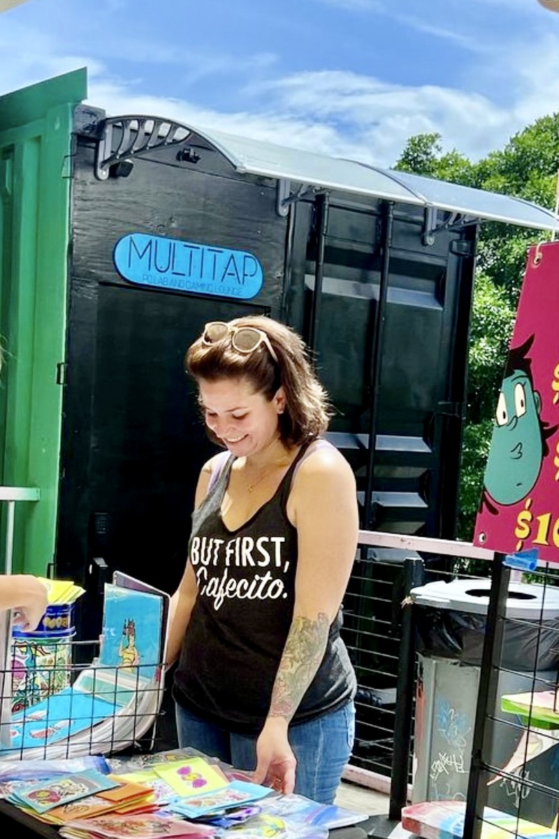 Silvia Maria standing beside a display of her colorful artwork at an outdoor market